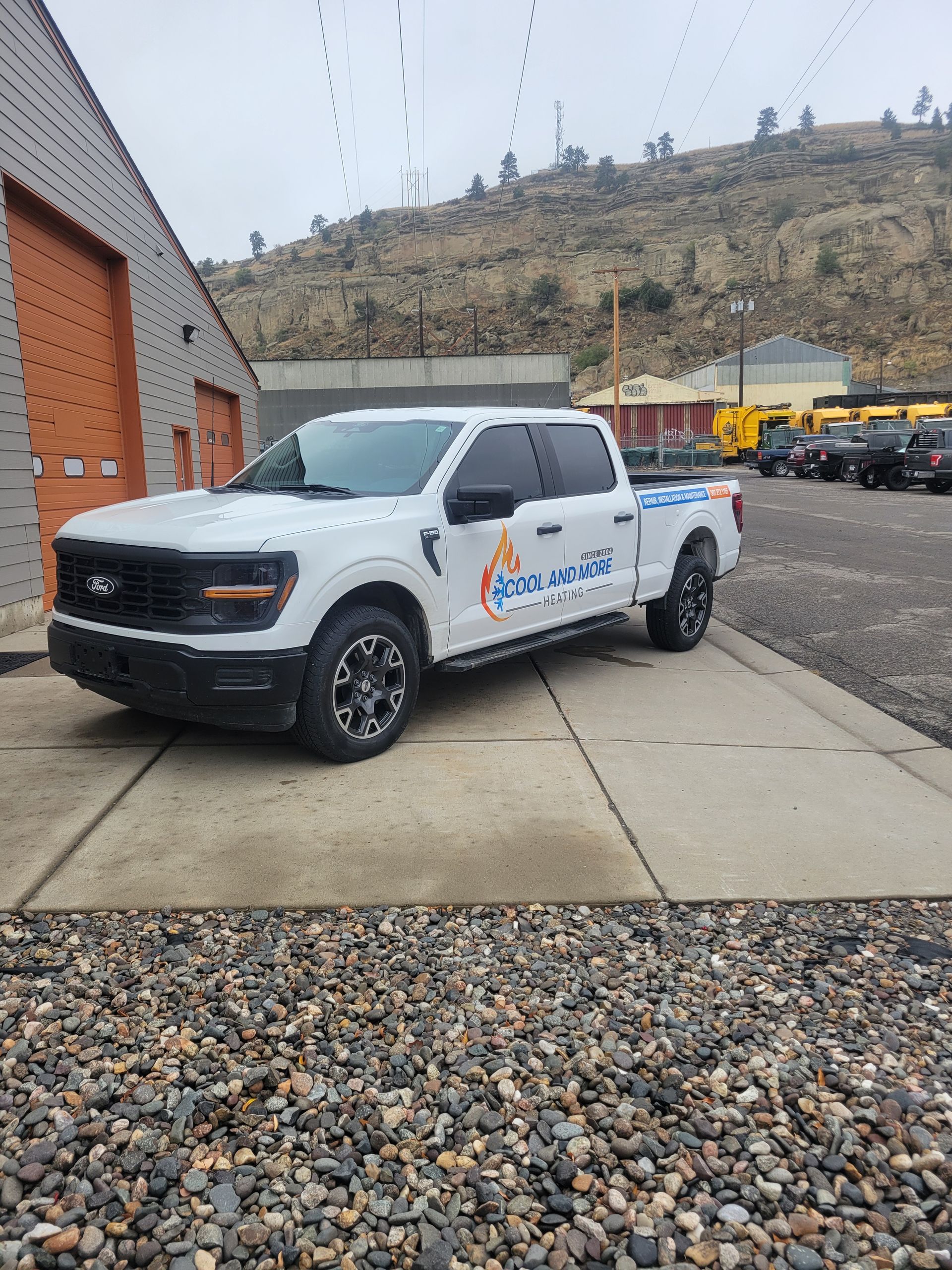 White pickup truck with company logo parked beside a brick building on a gravel lot, hills in background