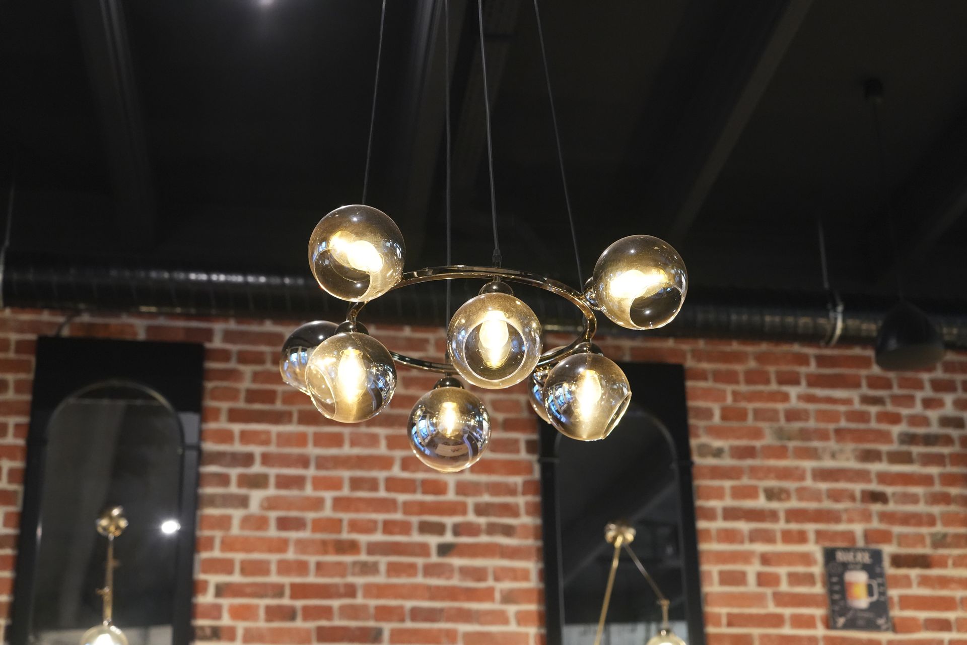 Modern chandelier with brass globe lights hanging in front of a brick wall and dark ceiling beams