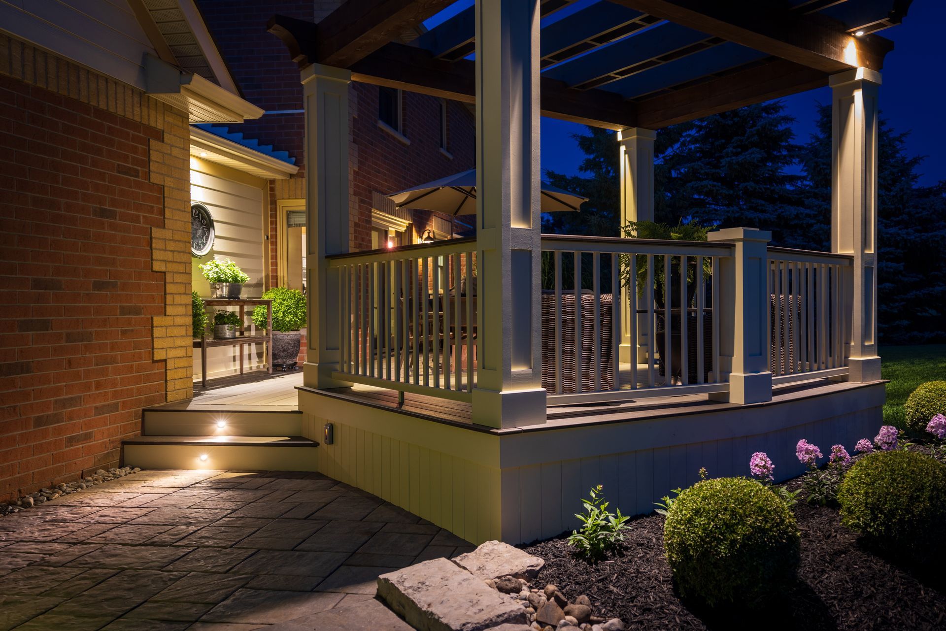 Lit patio with stone path, white railings, and warm porch lights beside a brick house at night