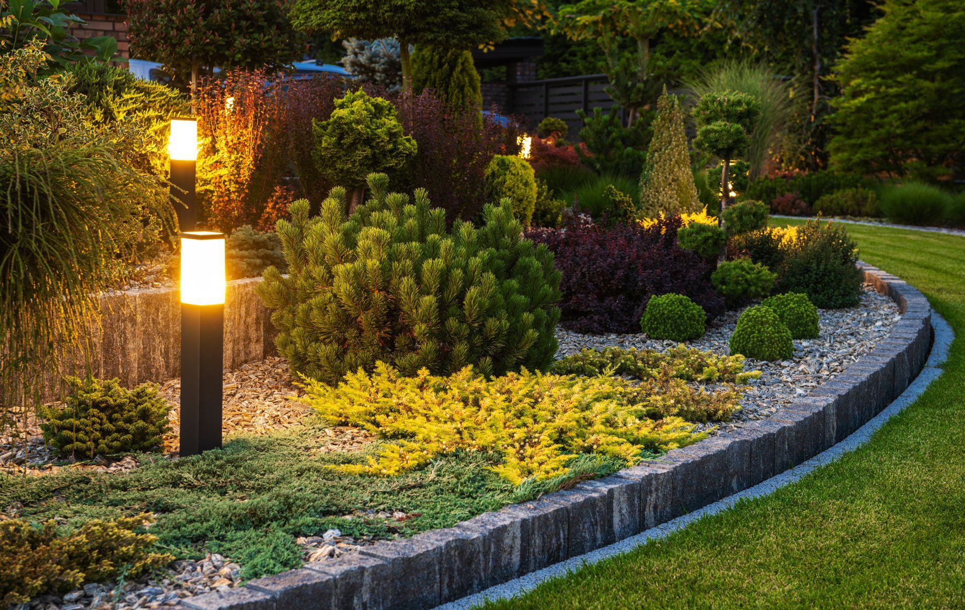 Curved garden border with glowing path lights, yellow flowers, shrubs, and a manicured lawn at dusk