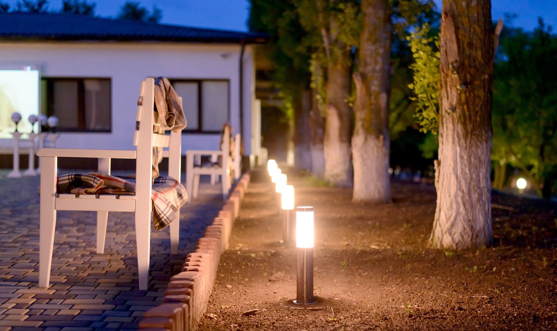 Lit garden path beside a white house with trees and glowing pathway lights at dusk