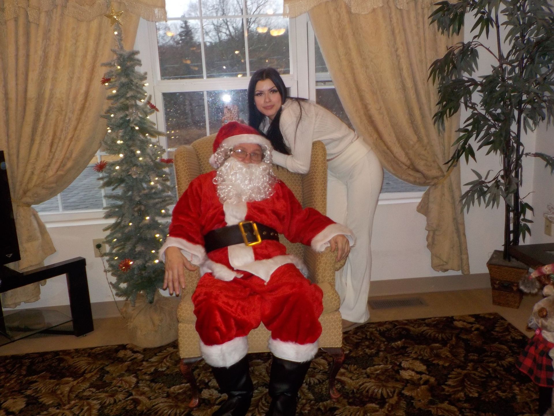 Santa Claus and woman pose near a window, Christmas tree, and plant. Santa wears red suit, woman white outfit.