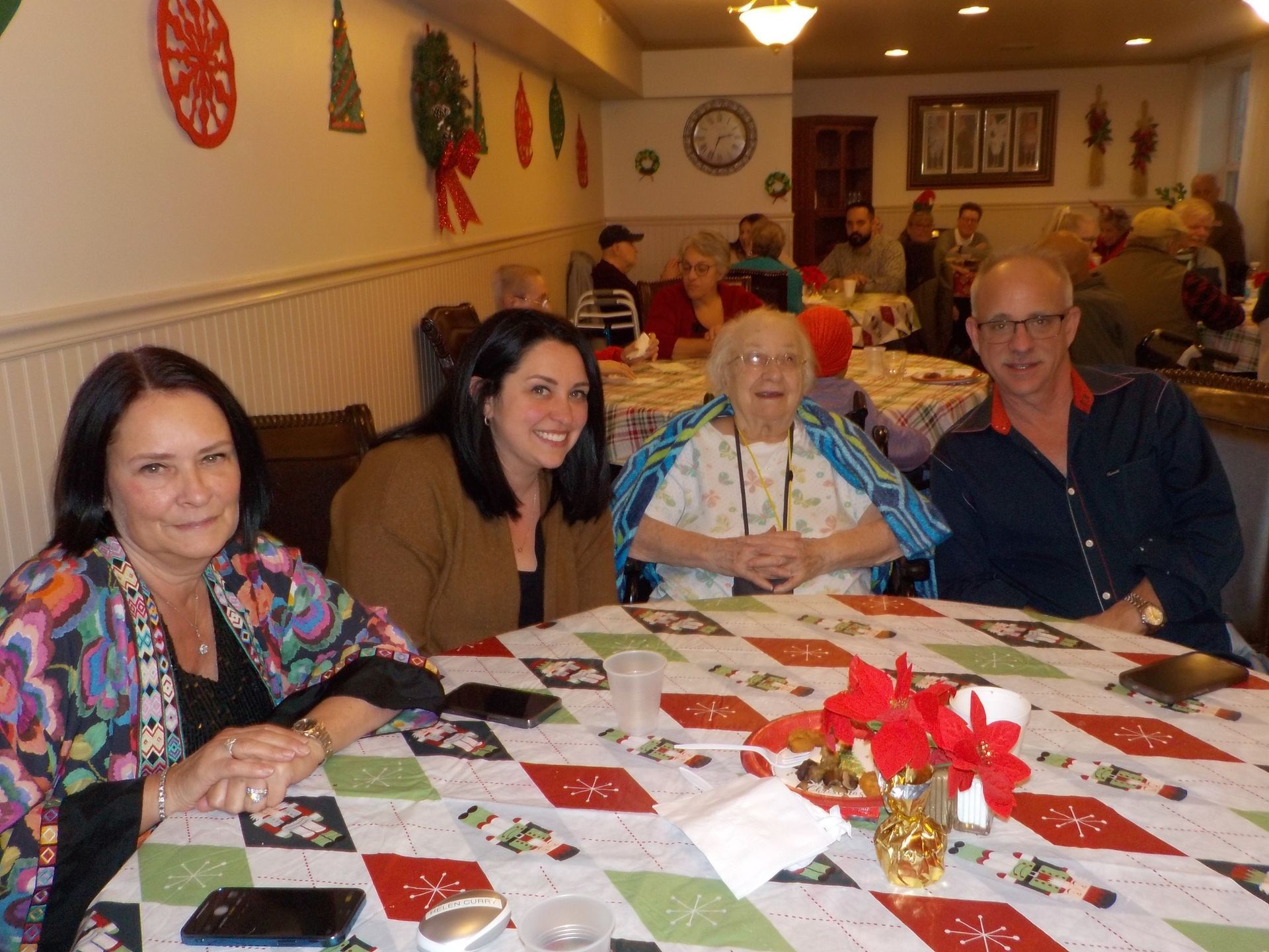 Four people seated at a table with a holiday tablecloth; others in the background; festive decorations.