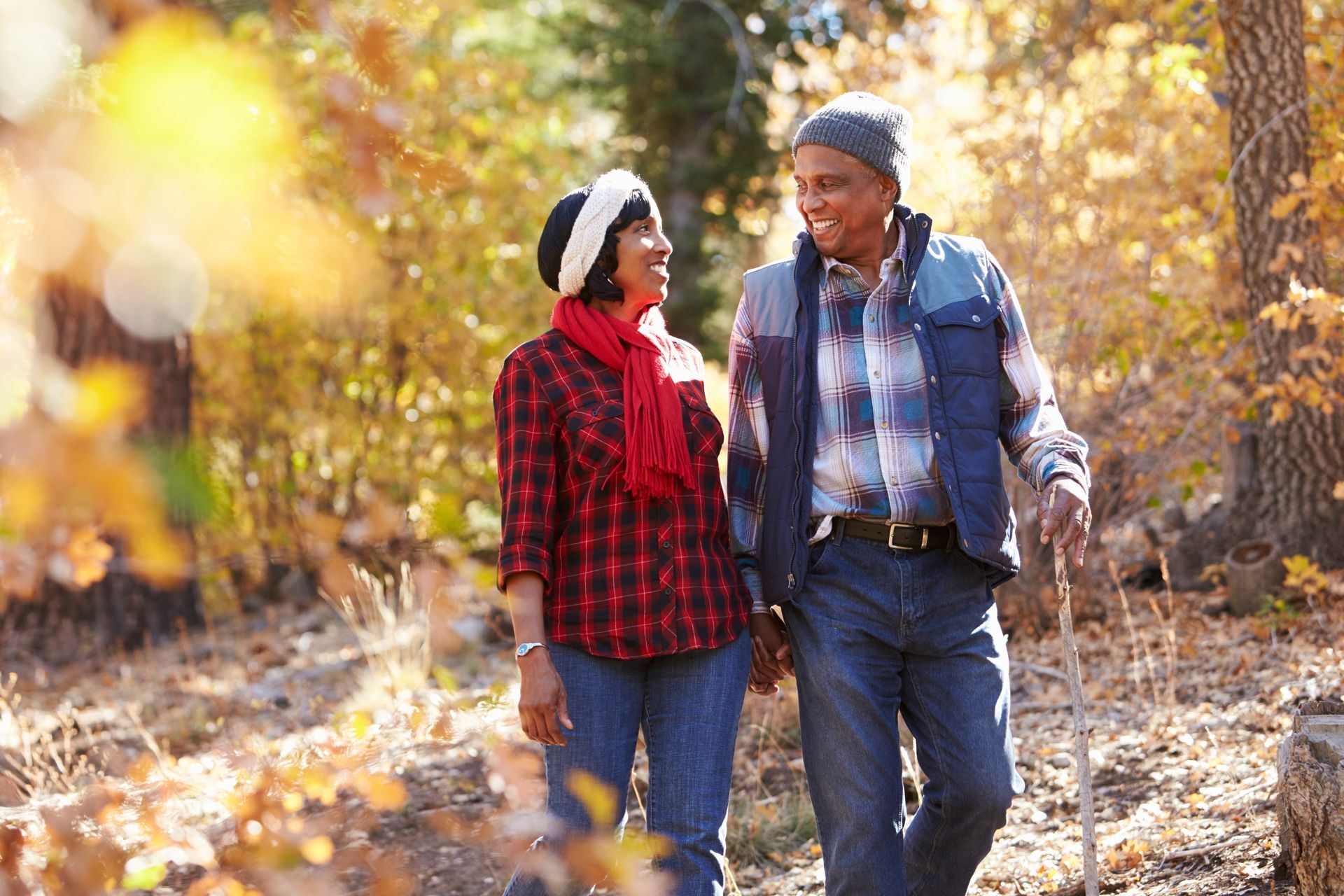 Two people walking together outdoors in an autumn forest, smiling and talking, with warm sunlight and fallen leaves.