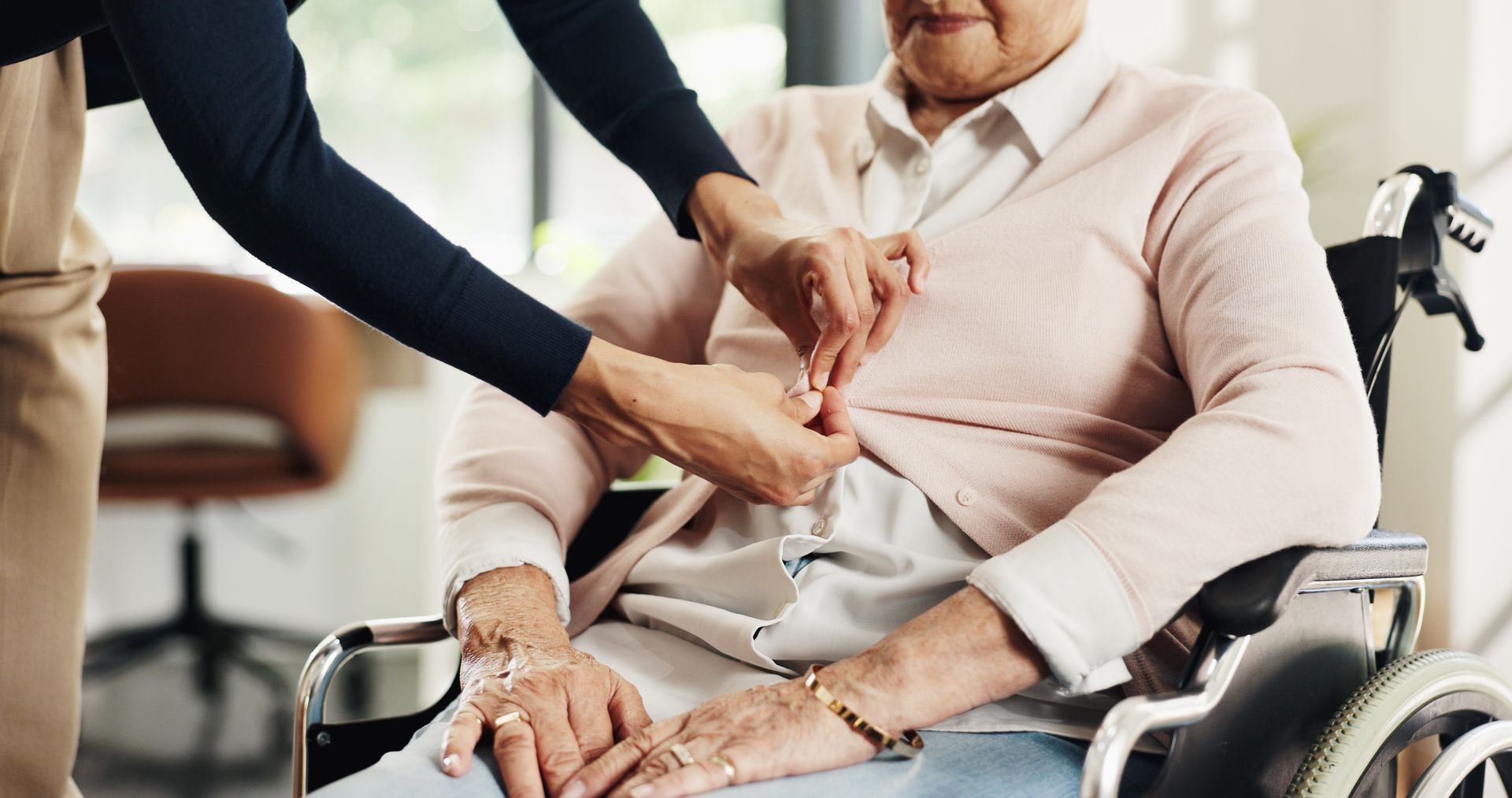 Caregiver fastening a seated patient’s shirt in a wheelchair, with a medical cane nearby.
