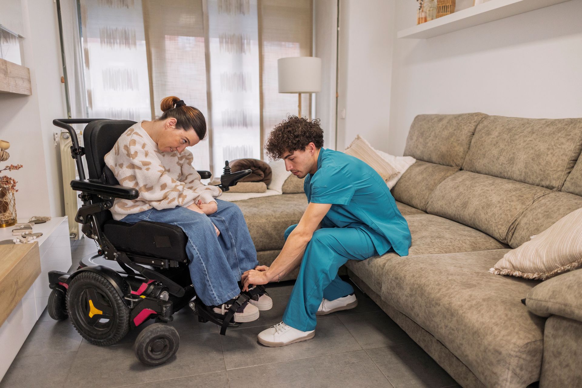 Caregiver kneels beside a woman in a wheelchair in a living room, helping adjust her foot placement.