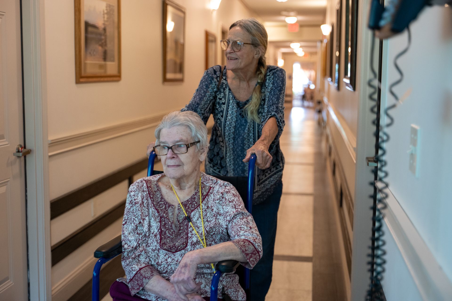 Two women in a hallway, one seated in a wheelchair while another pushes from behind.
