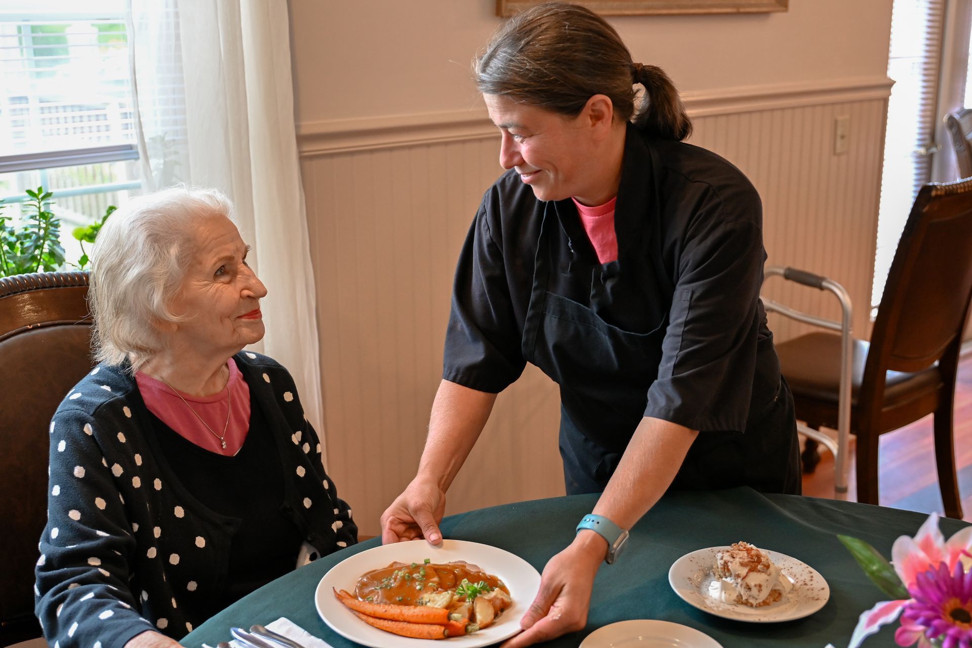 Woman serving a plate of food to an elderly woman at a table.