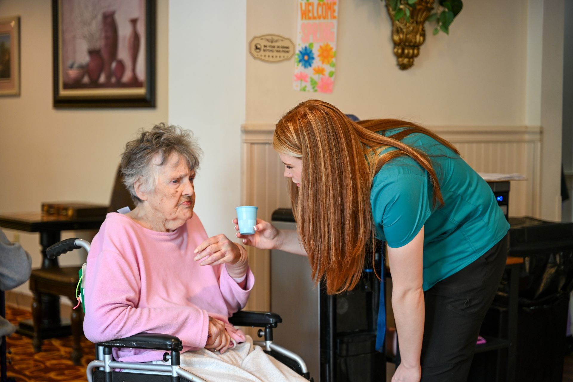 A caregiver offers medication to an elderly woman in a wheelchair.