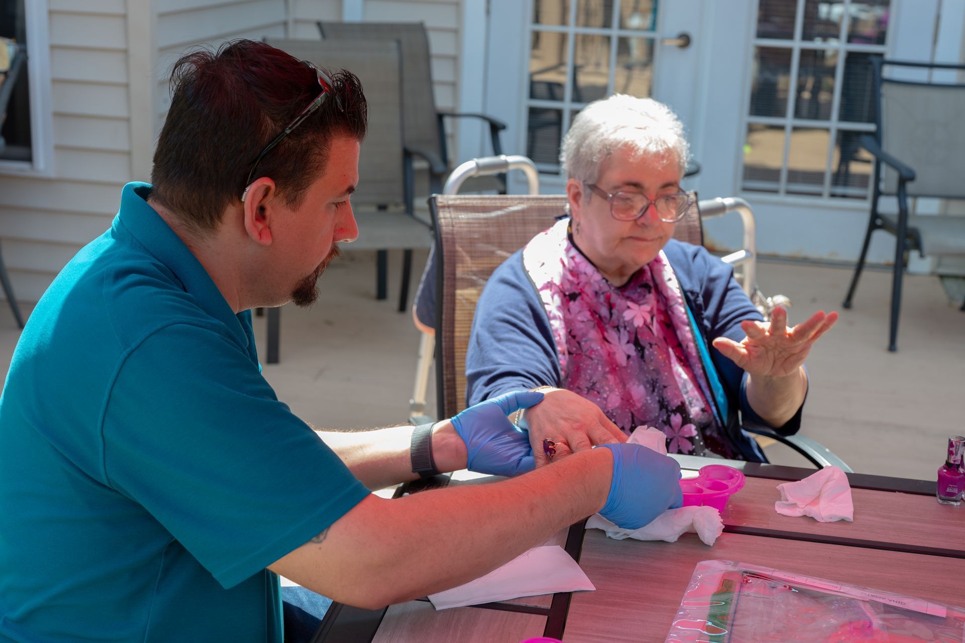 Two people sit at a table outdoors, working with craft supplies and pink paper.