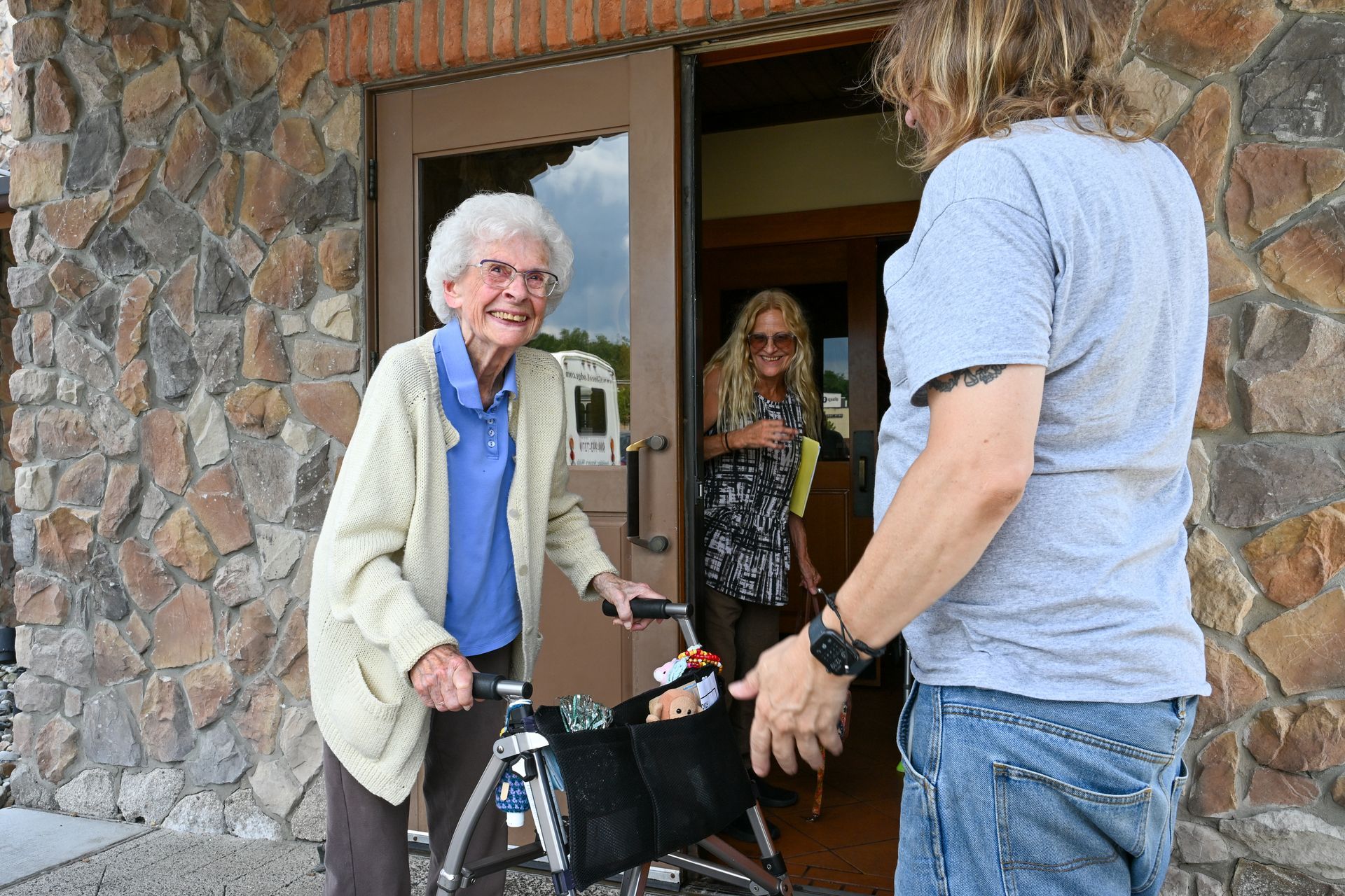Elderly woman with walker, being helped out of a building by a person, another woman in the doorway.