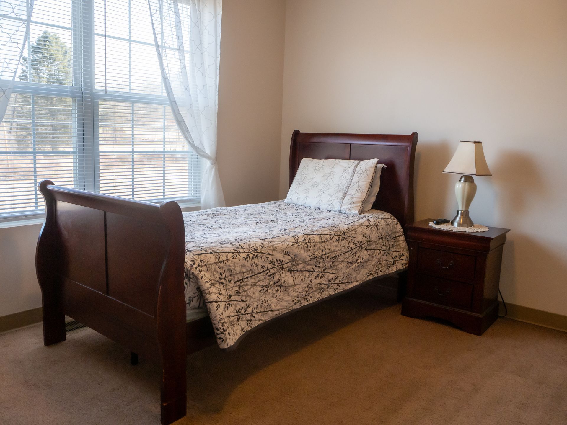 Bedroom with a dark wood bed, patterned bedding, nightstand lamp, and a large window with sheer curtains.