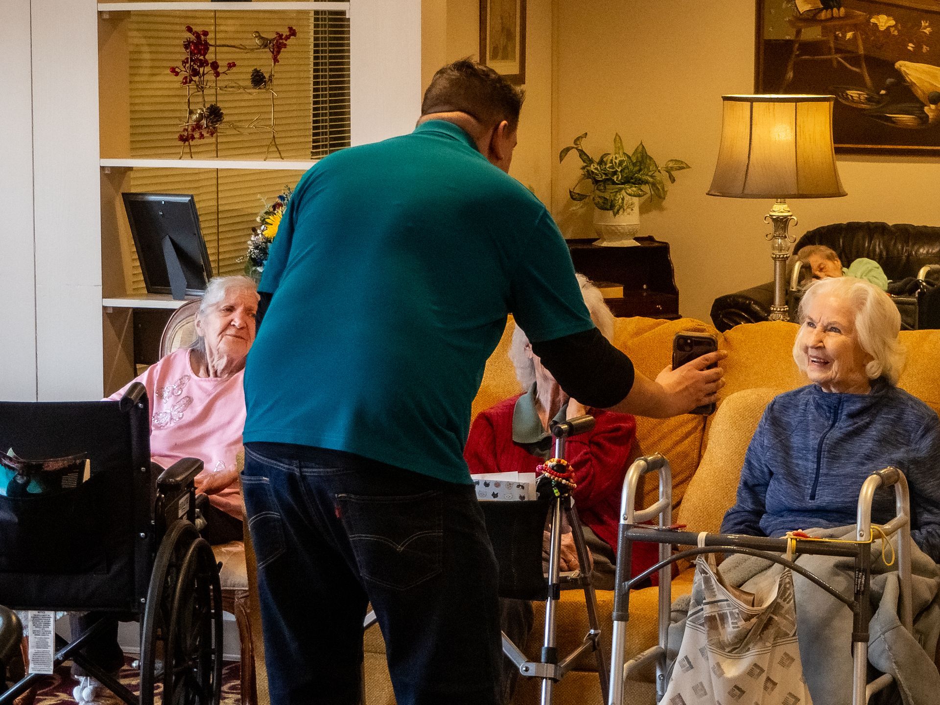 Man taking photo with elderly women in a living room, one in a wheelchair.