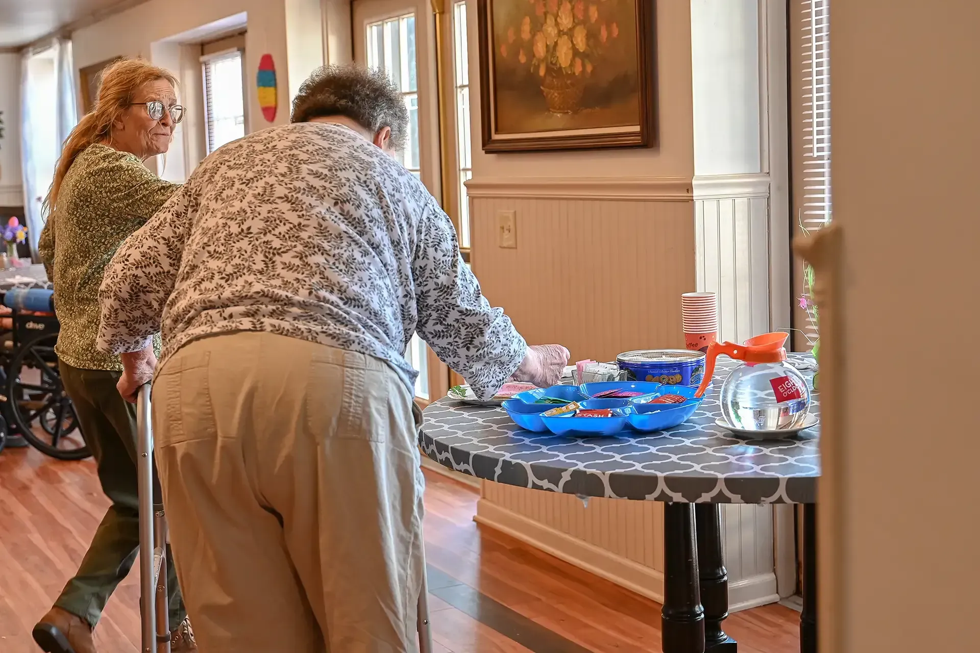 Two elderly women near a table. One uses a walker. The table has supplies, a painting on the wall.
