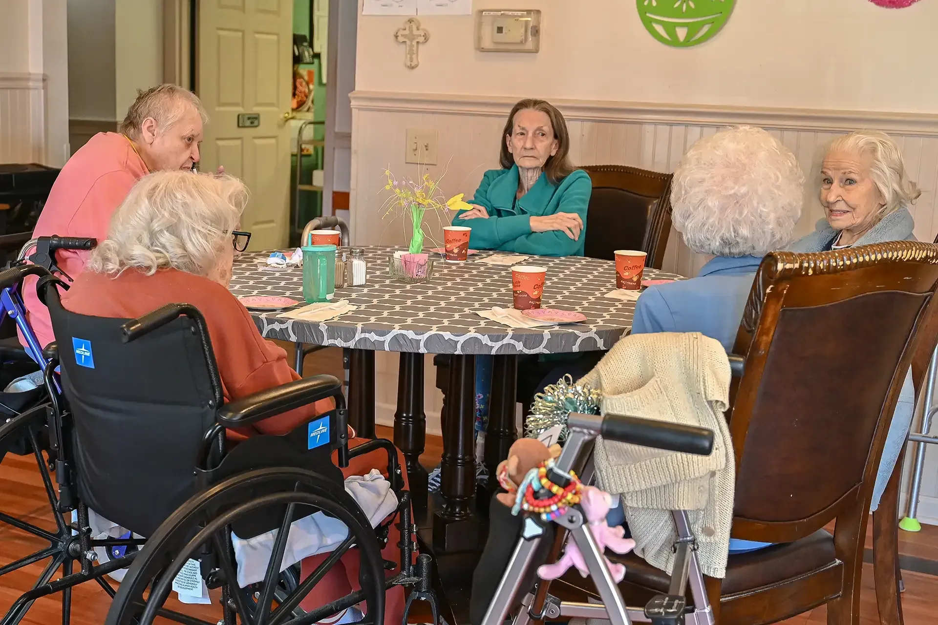 Group of seniors and a woman seated at a round table, some in wheelchairs, conversing indoors.