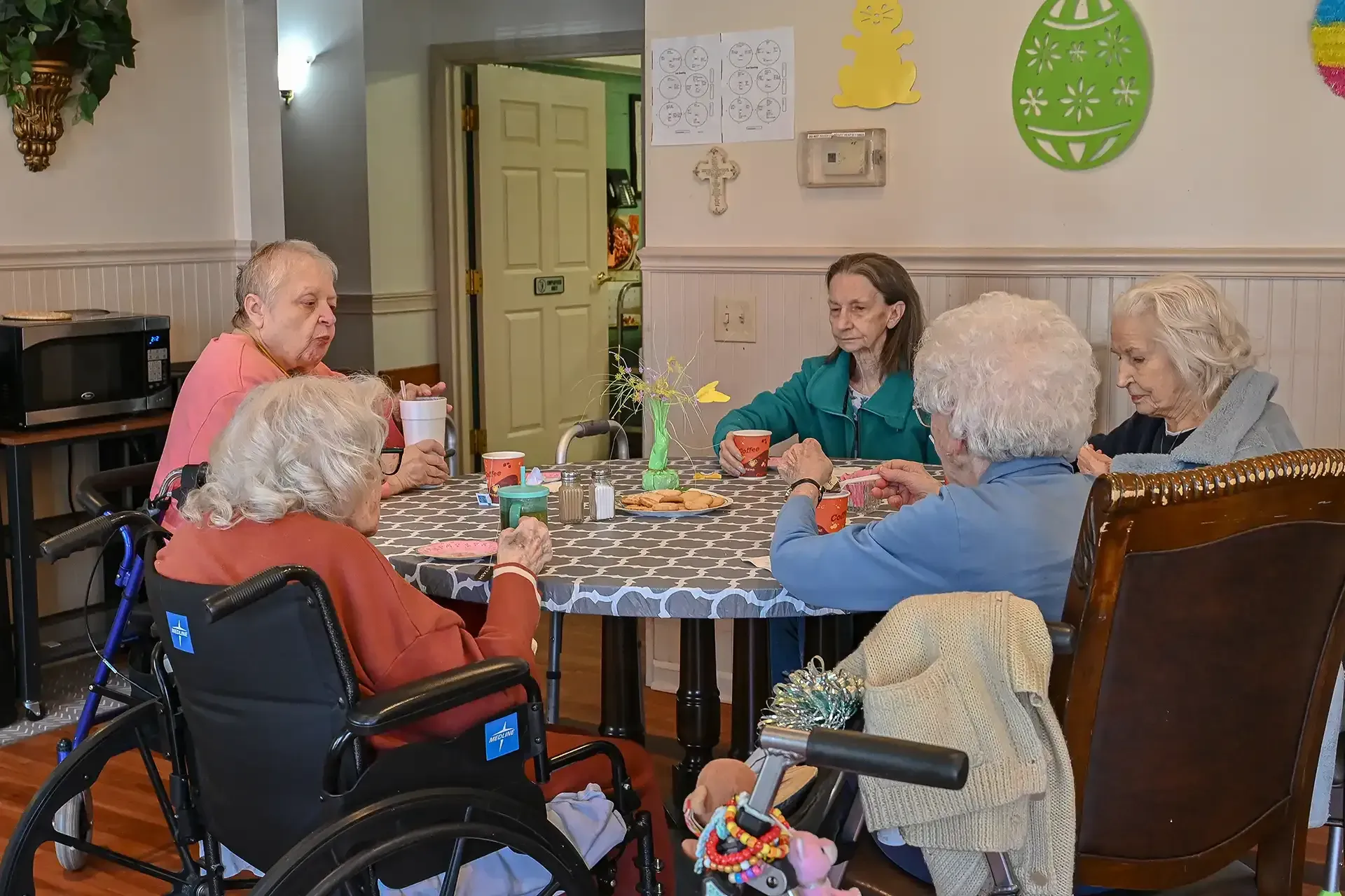 Five people at a table in a room. Some are in wheelchairs. They appear to be talking and holding cups.