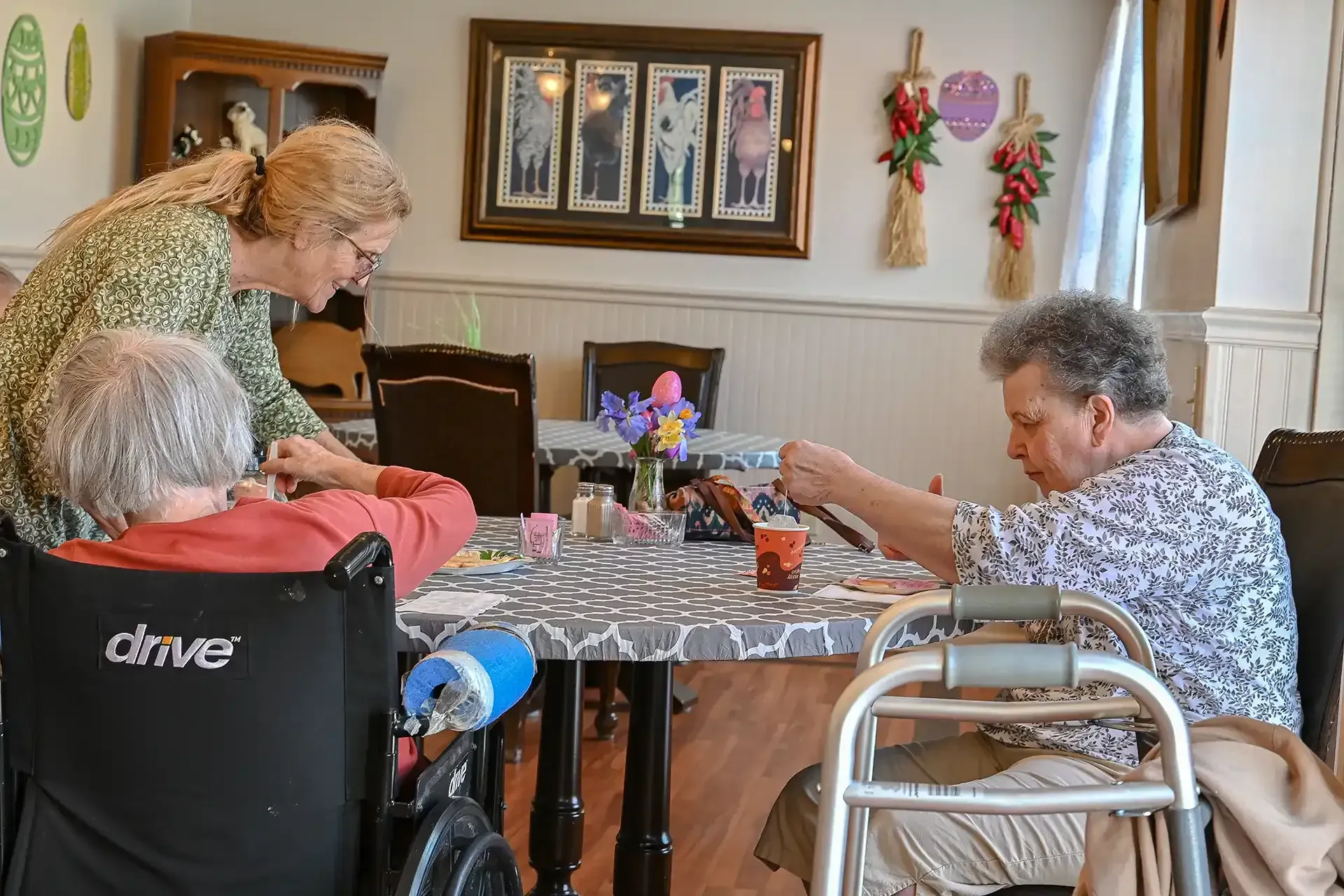 Woman assisting two seniors crafting at a table; sunny room.