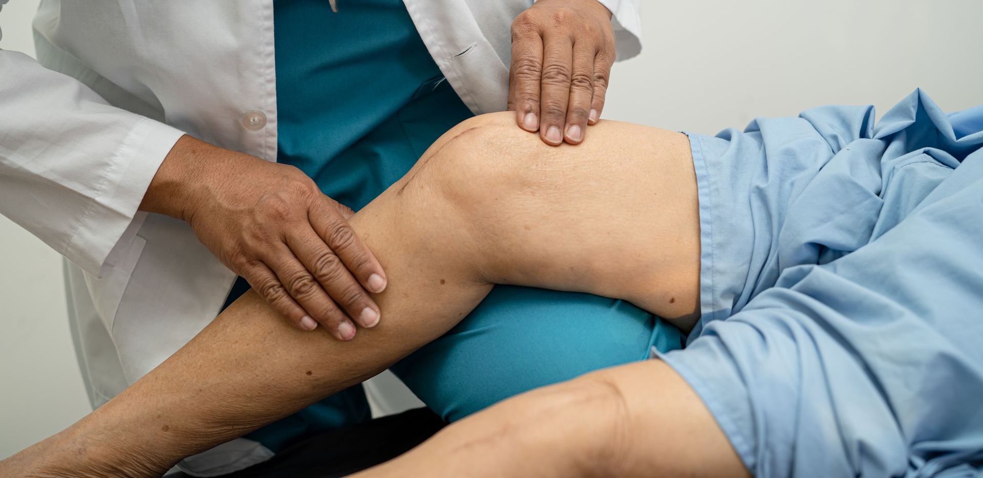 Pflegekraft im OP-Kittel hilft einem älteren Patienten im Bett und untersucht sein Knie. Zimmer mit Rollstuhl.