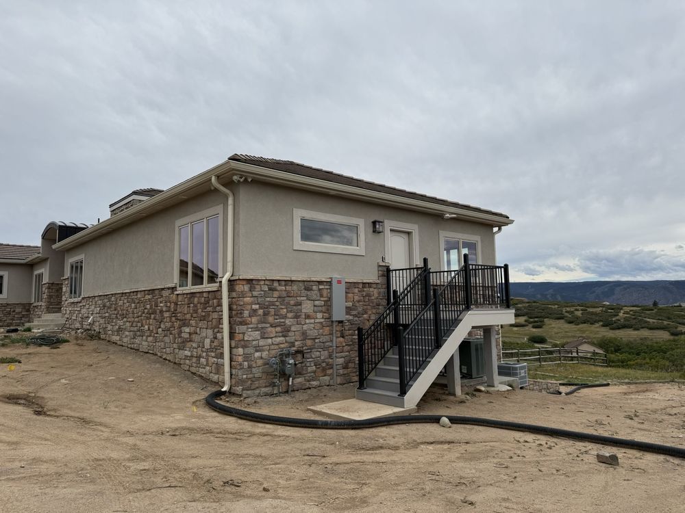 Modern single-story house with stone siding and exterior stairs on a dirt lot under a cloudy sky