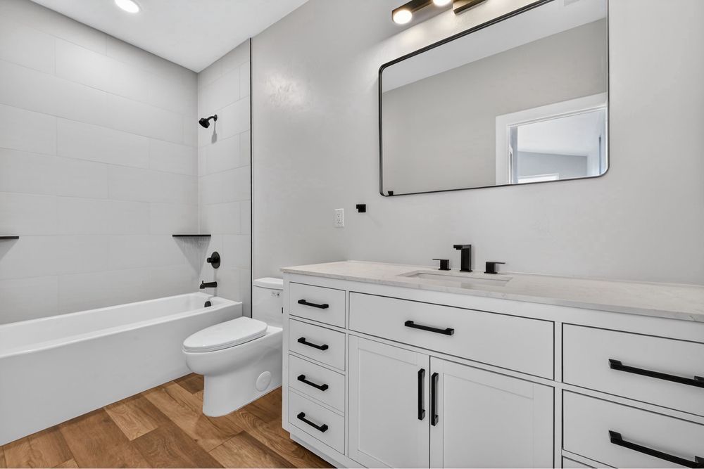 Bright white bathroom with bathtub, toilet, and double-sink vanity with mirror and wood floor