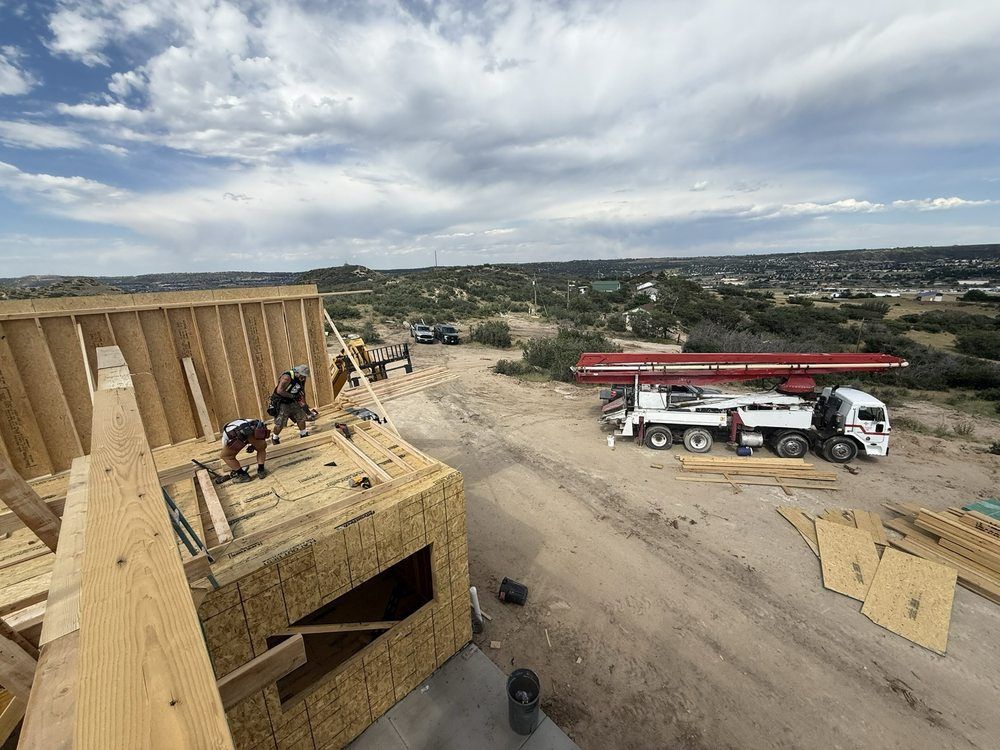 Workers on a wooden structure at a construction site, with trucks and open landscape in the background.