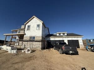 Two-story white house under construction with a black pickup truck parked on a dirt lot under a clear blue sky