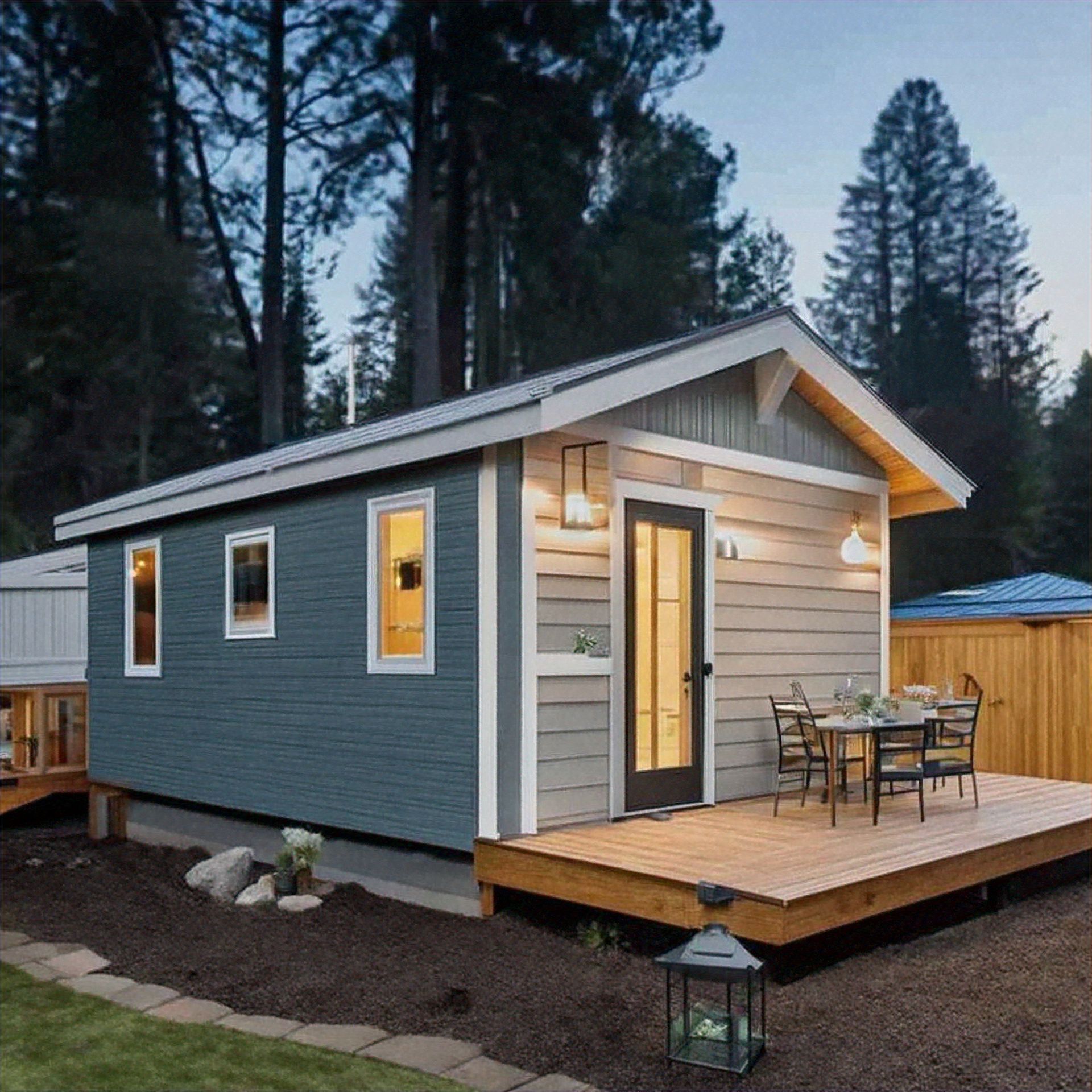 Small gray cabin with a wooden porch and outdoor table, set among tall trees at dusk.