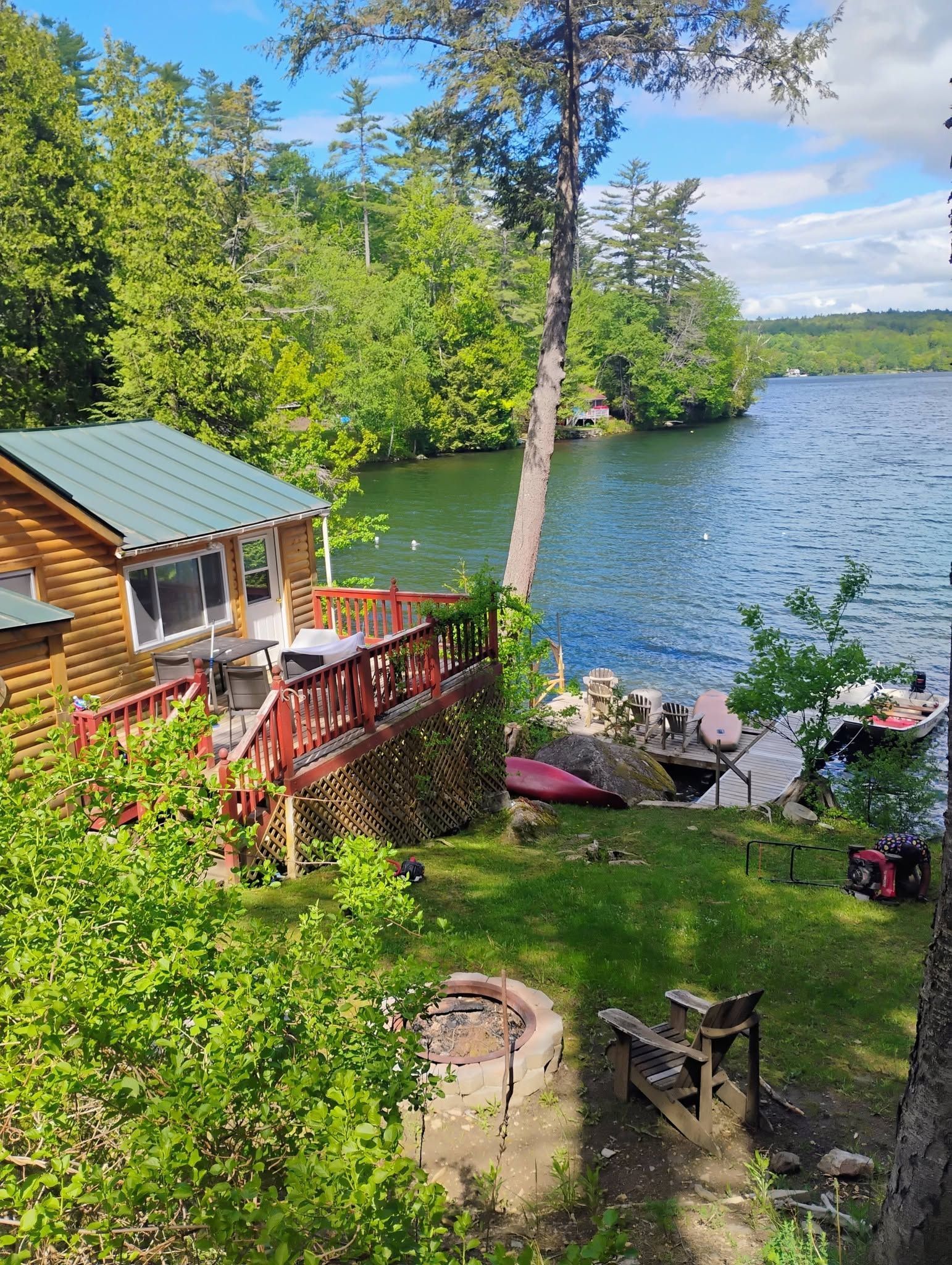Lakeside cabin with red deck, green trees, fire pit, and small boats by a calm blue lake.