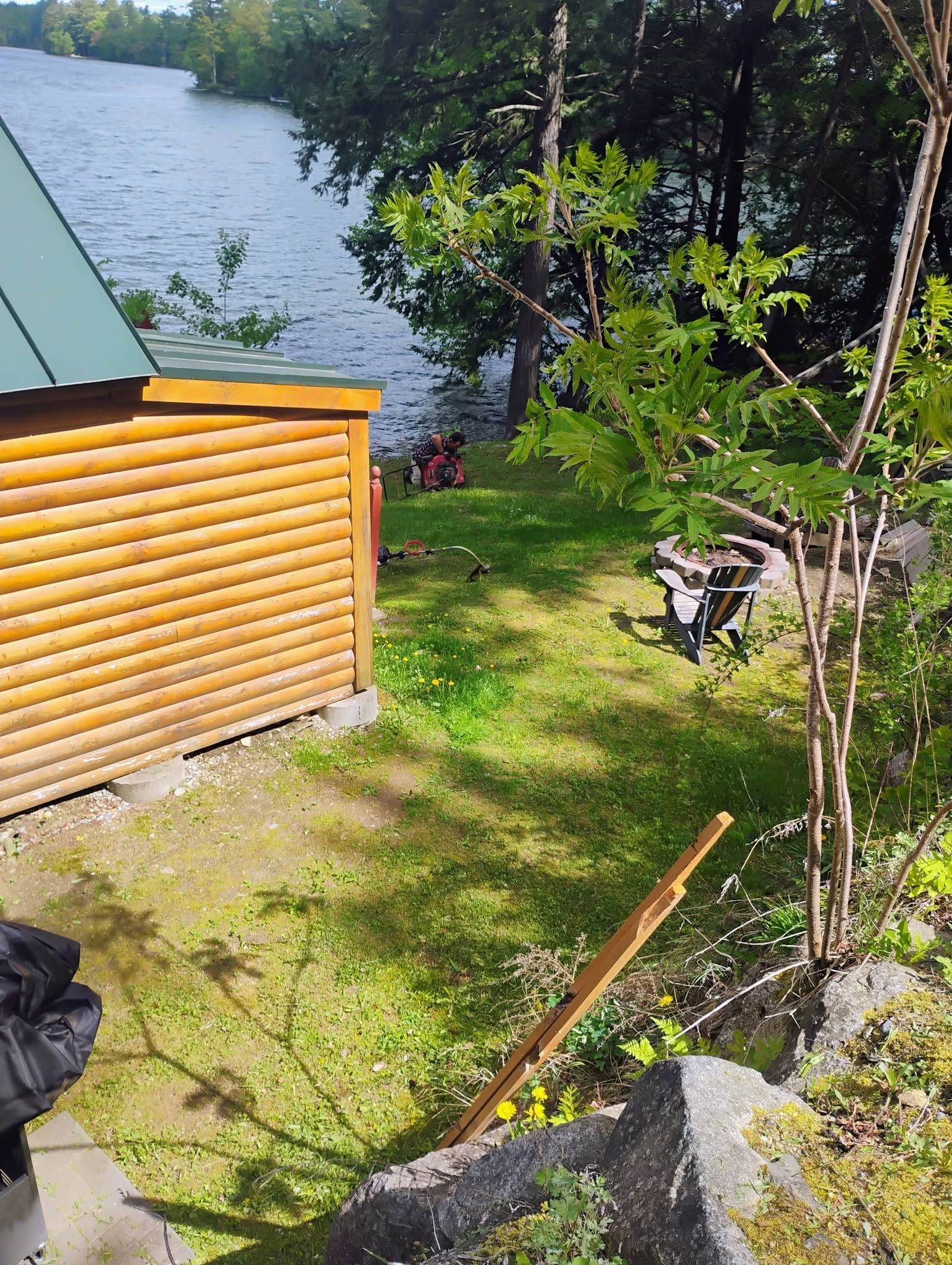 Wooden cabin beside a lake, with grassy yard, trees, and rocks in bright sunlight.