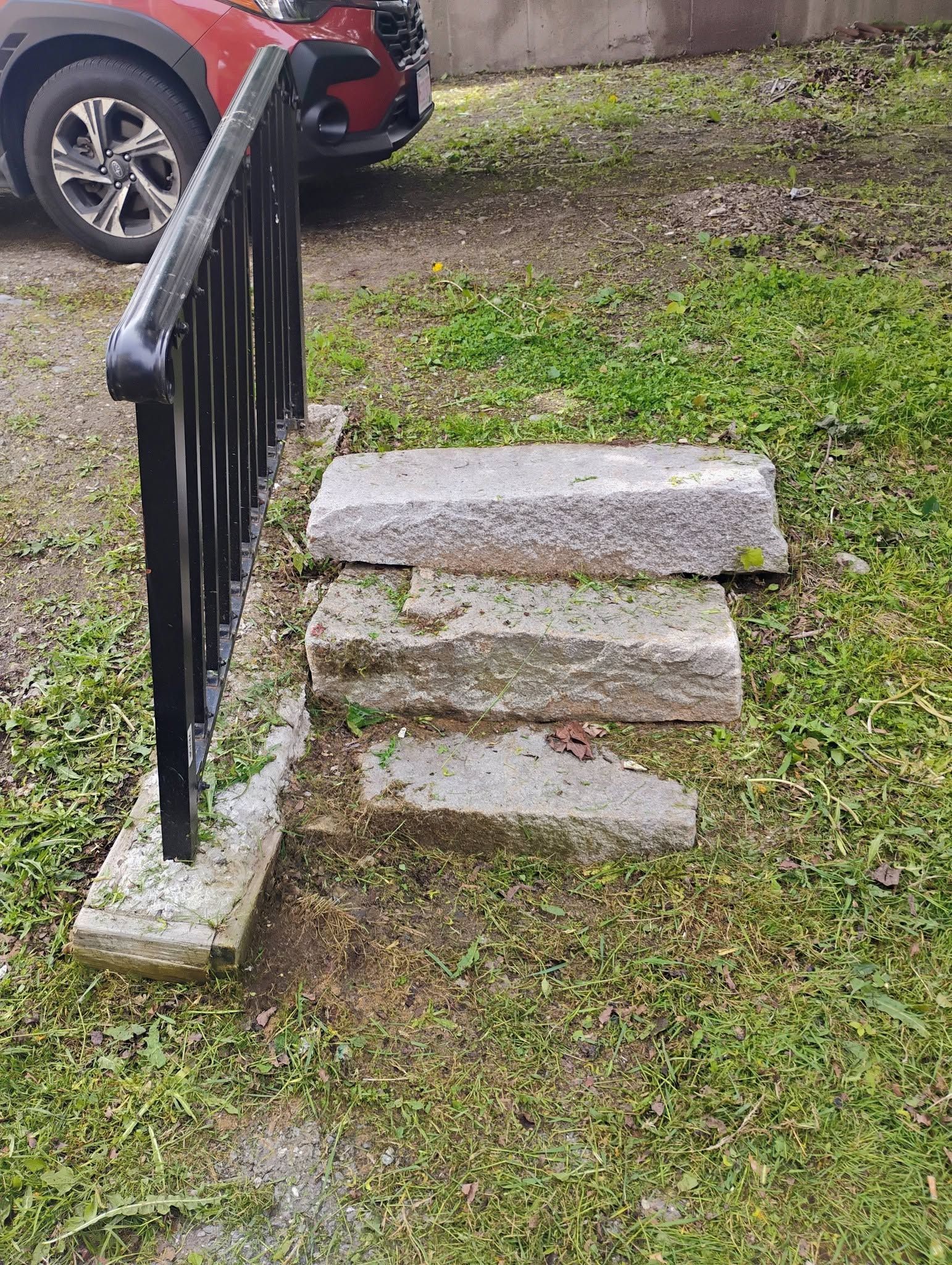 Three uneven stone steps beside a black railing, leading up from a grassy yard near a parked red car.
