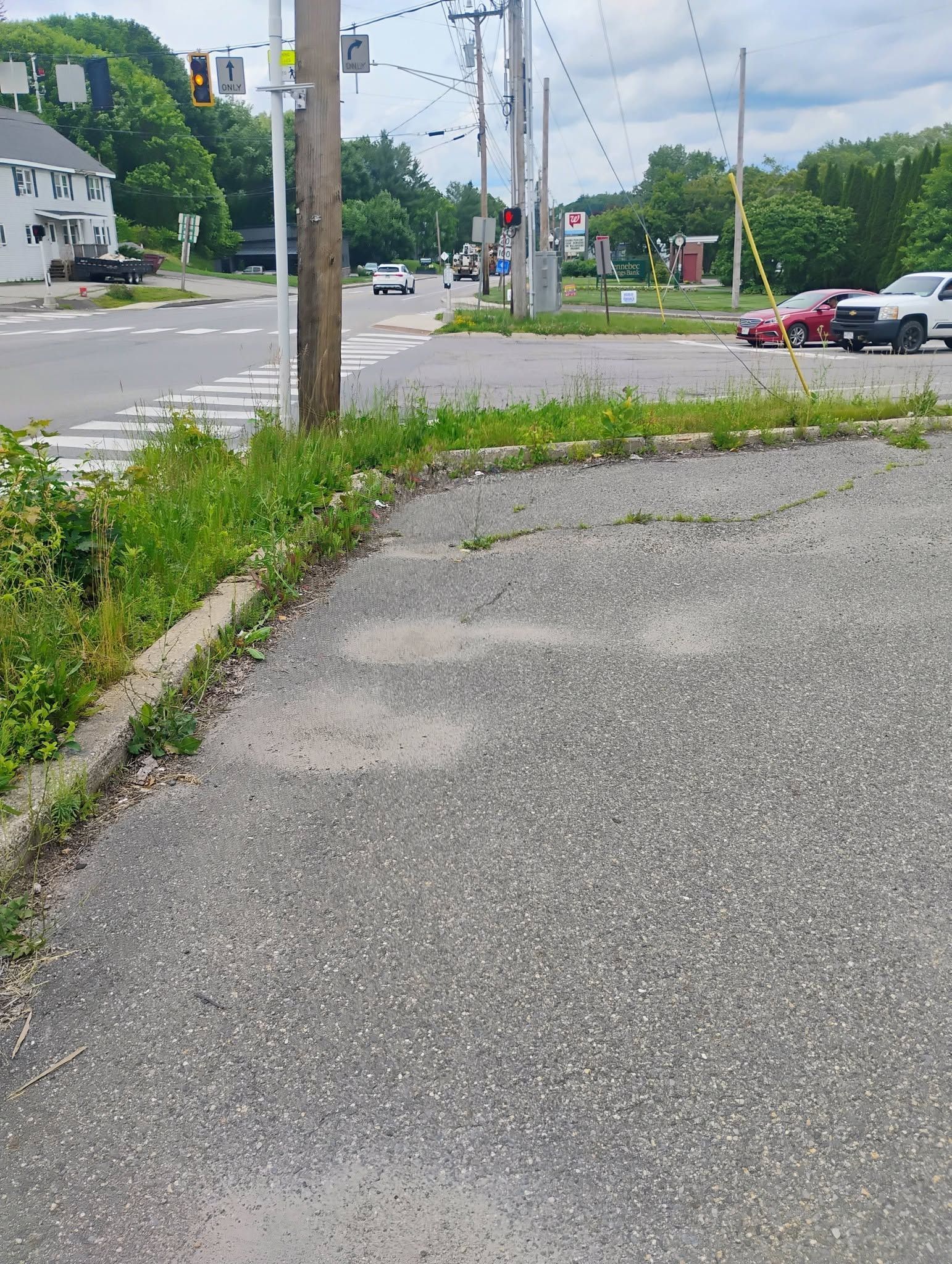 Cracked sidewalk beside a grassy roadside near an intersection with utility poles and parked cars.