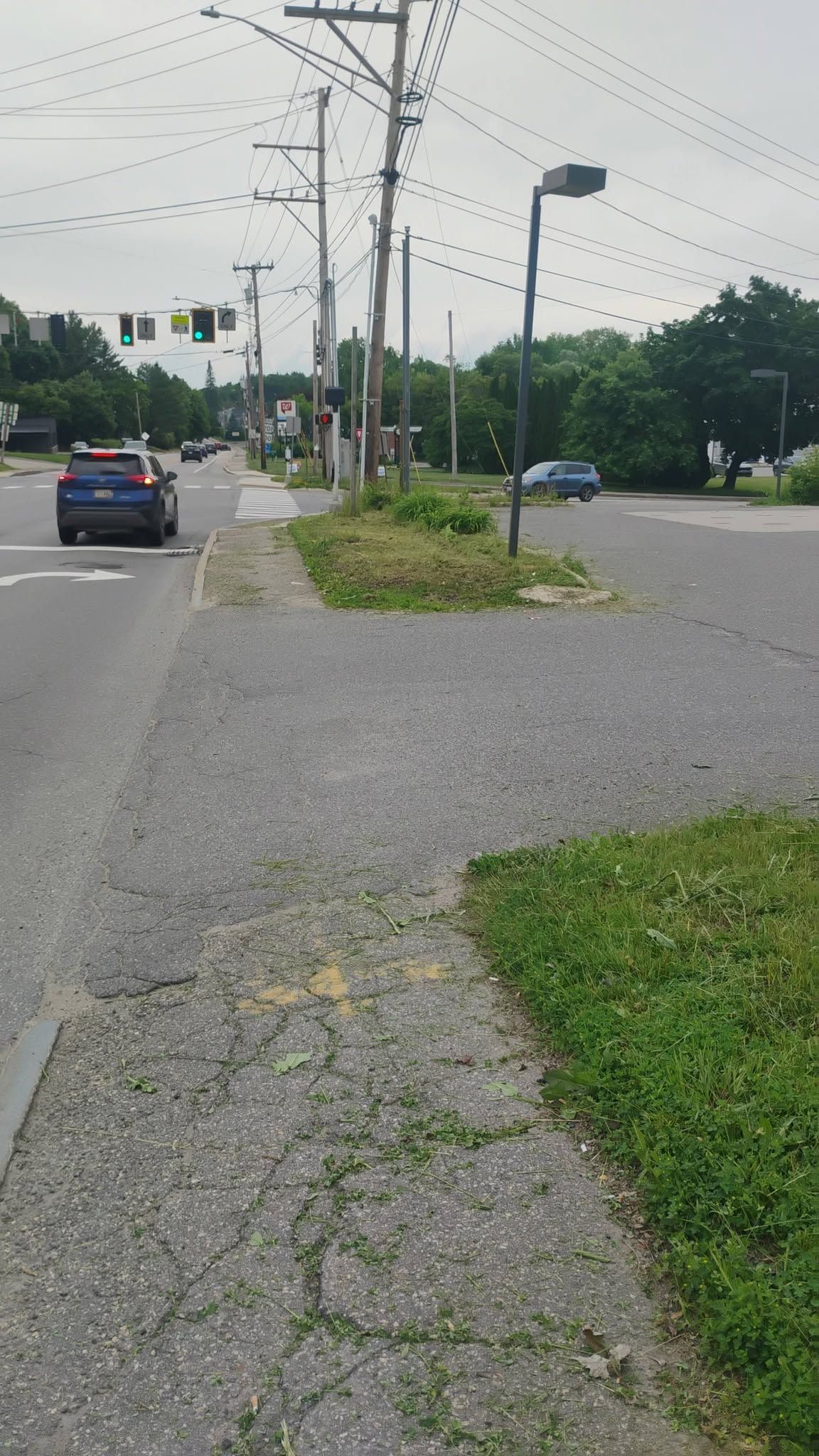 Sidewalk beside a road intersection with cars, utility poles, and grassy median under cloudy sky