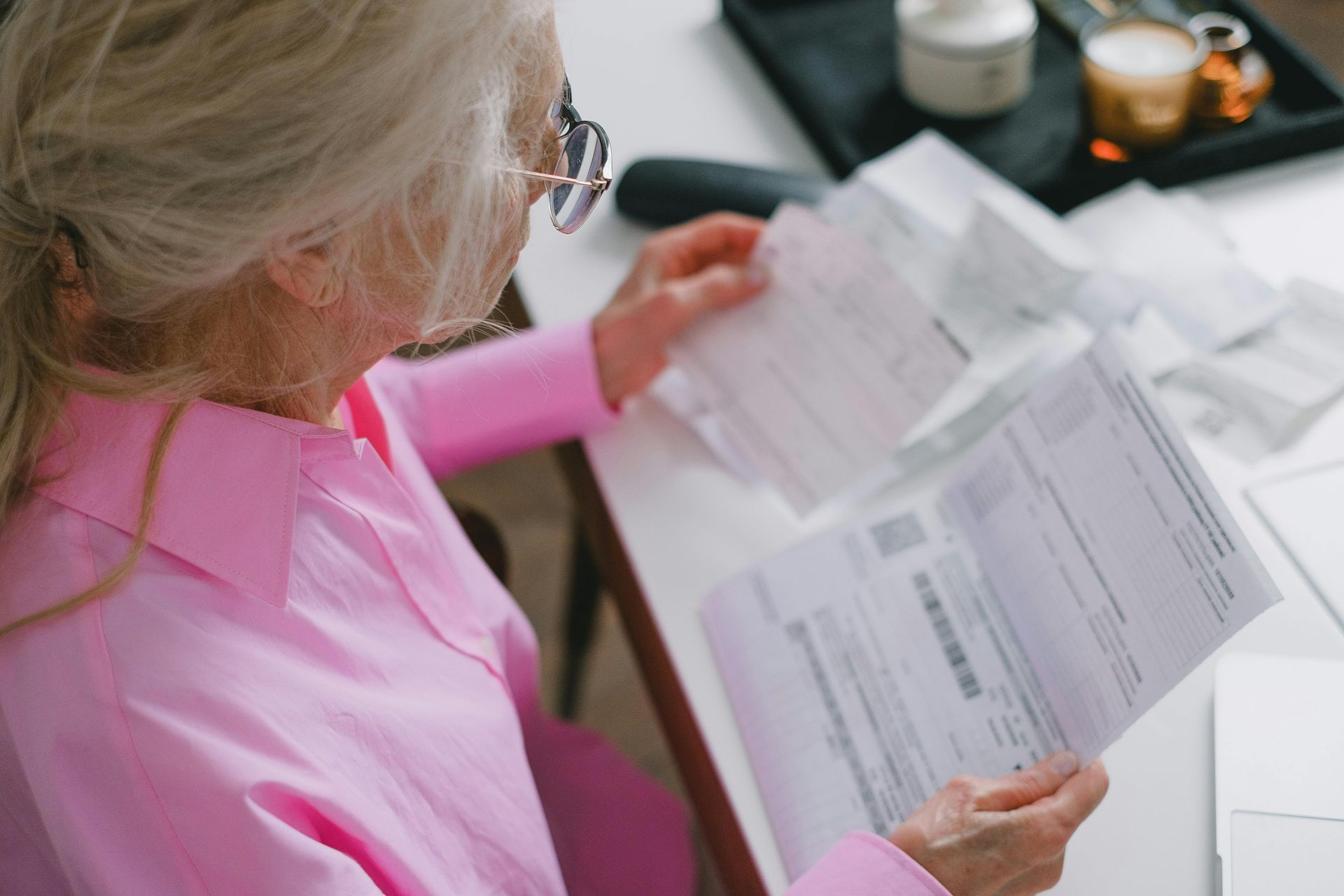 Person in a pink shirt reviewing paperwork at a desk with a laptop and coffee cups