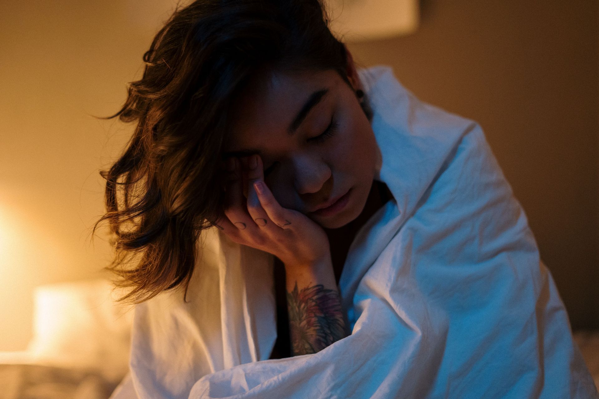 Woman sitting up in bed with her head resting on her hand with eyes closed, appearing exhausted