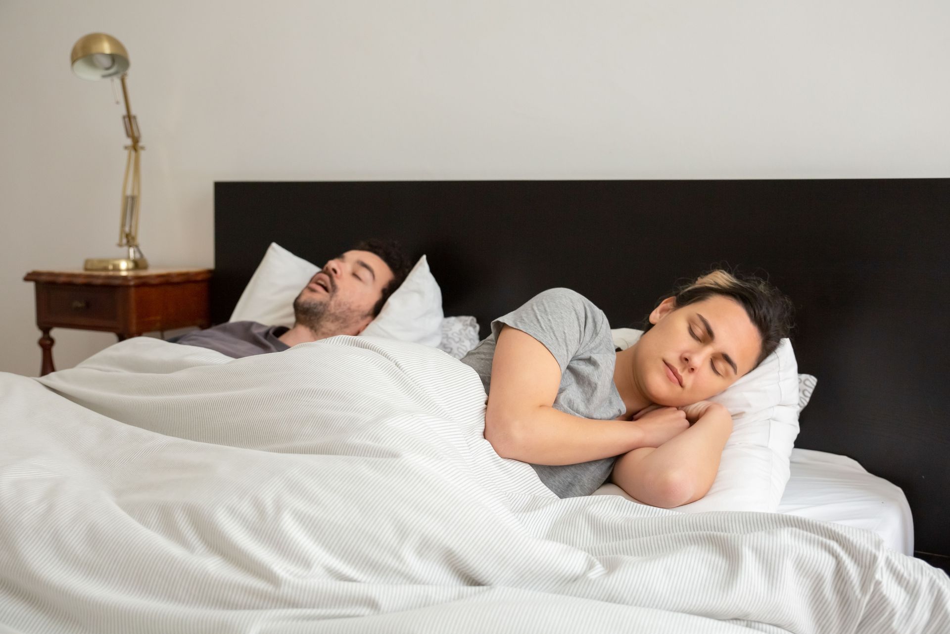 Man and woman lying in bed with the man appearing to be snoring and the woman trying to go to sleep