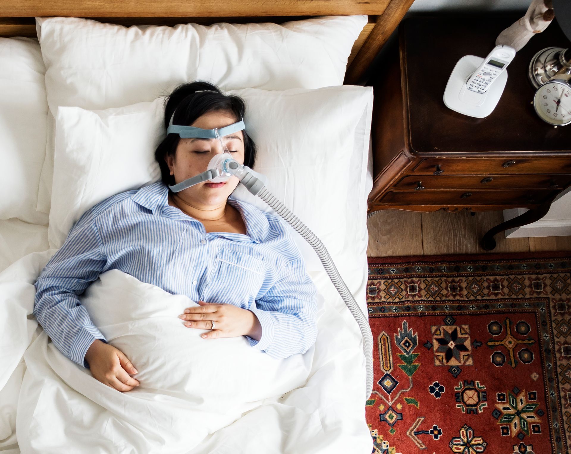 Person lying in bed with an oxygen mask, covered by a white blanket beside a wooden nightstand.