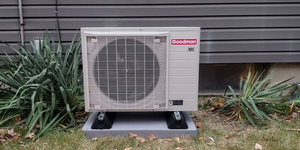 Outdoor air conditioning unit on a concrete pad beside a house wall and shrubs