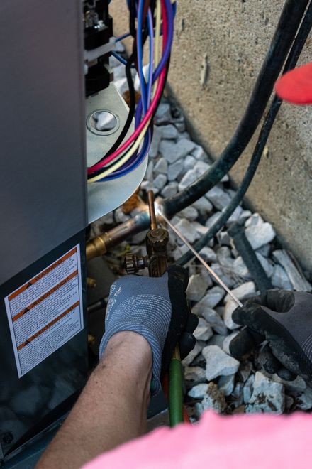 Technician adjusting brass HVAC valve near an outdoor unit, with gloves and gravel ground visible
