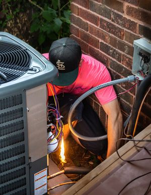 Worker in pink shirt welding beside a house, with sparks flying near an air conditioner unit.