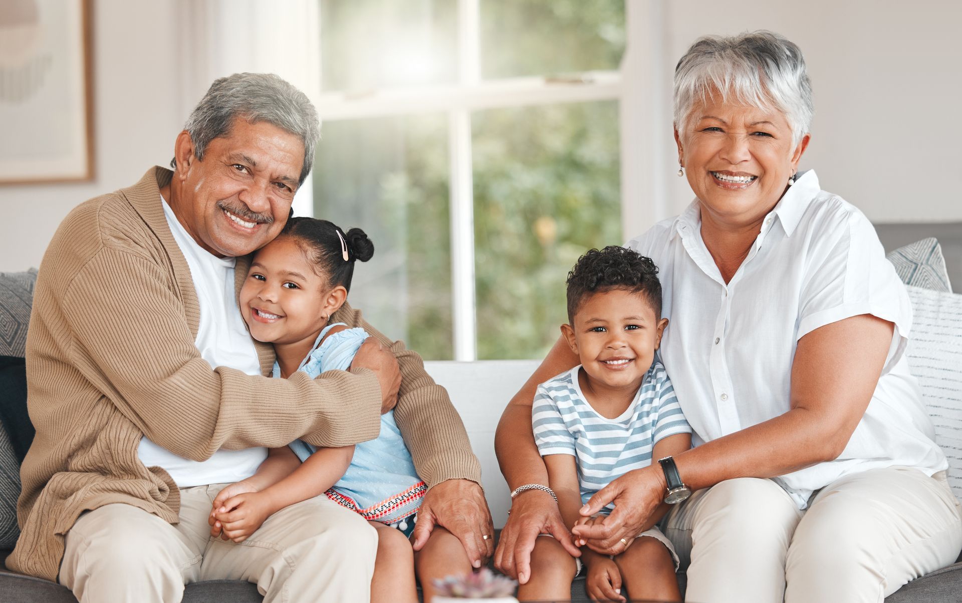 A smiling couple sits on a sofa with two young children, all embracing in a bright, indoor living room setting.