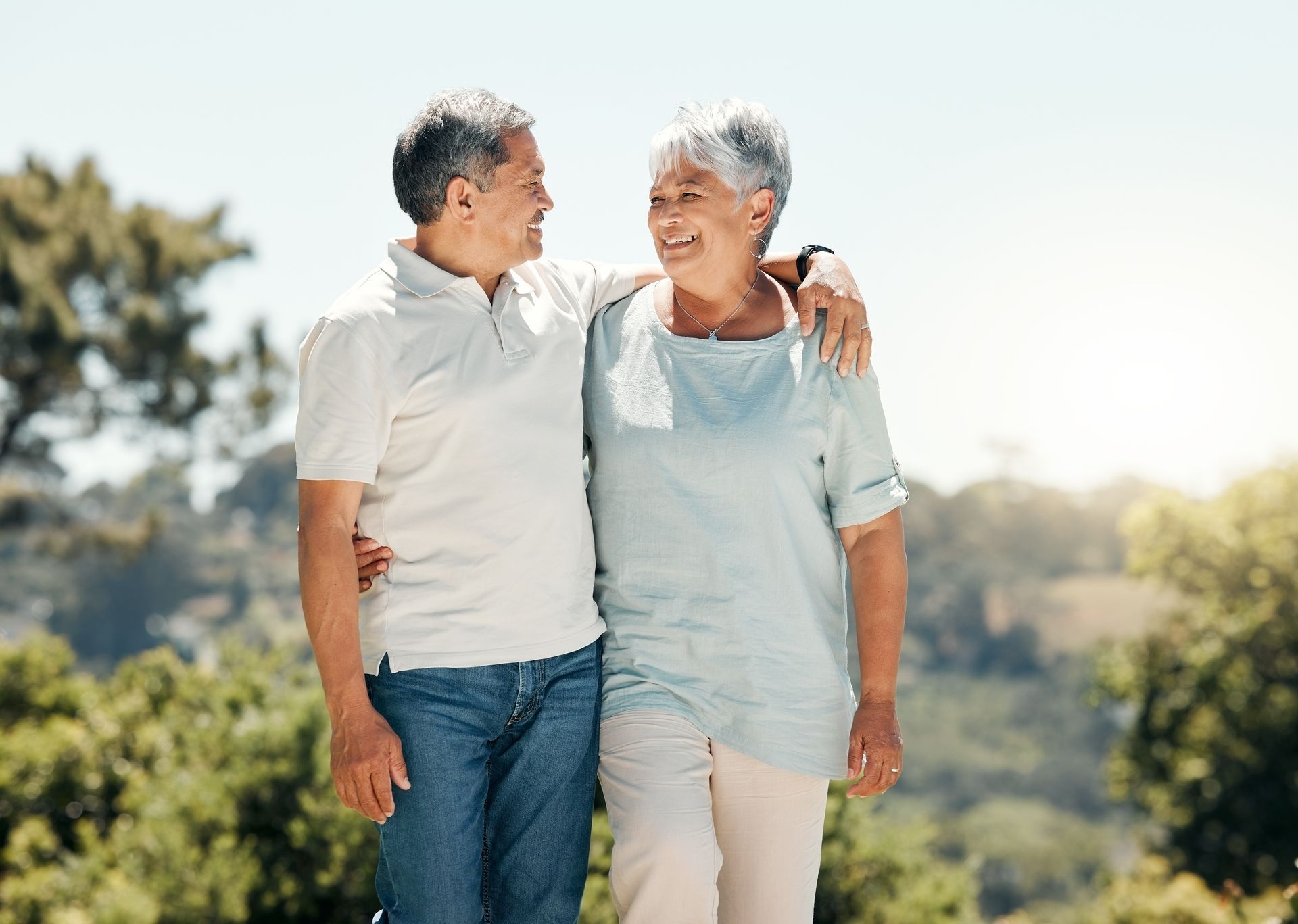 Two people walking side by side outdoors, smiling at each other with their arms around one another.