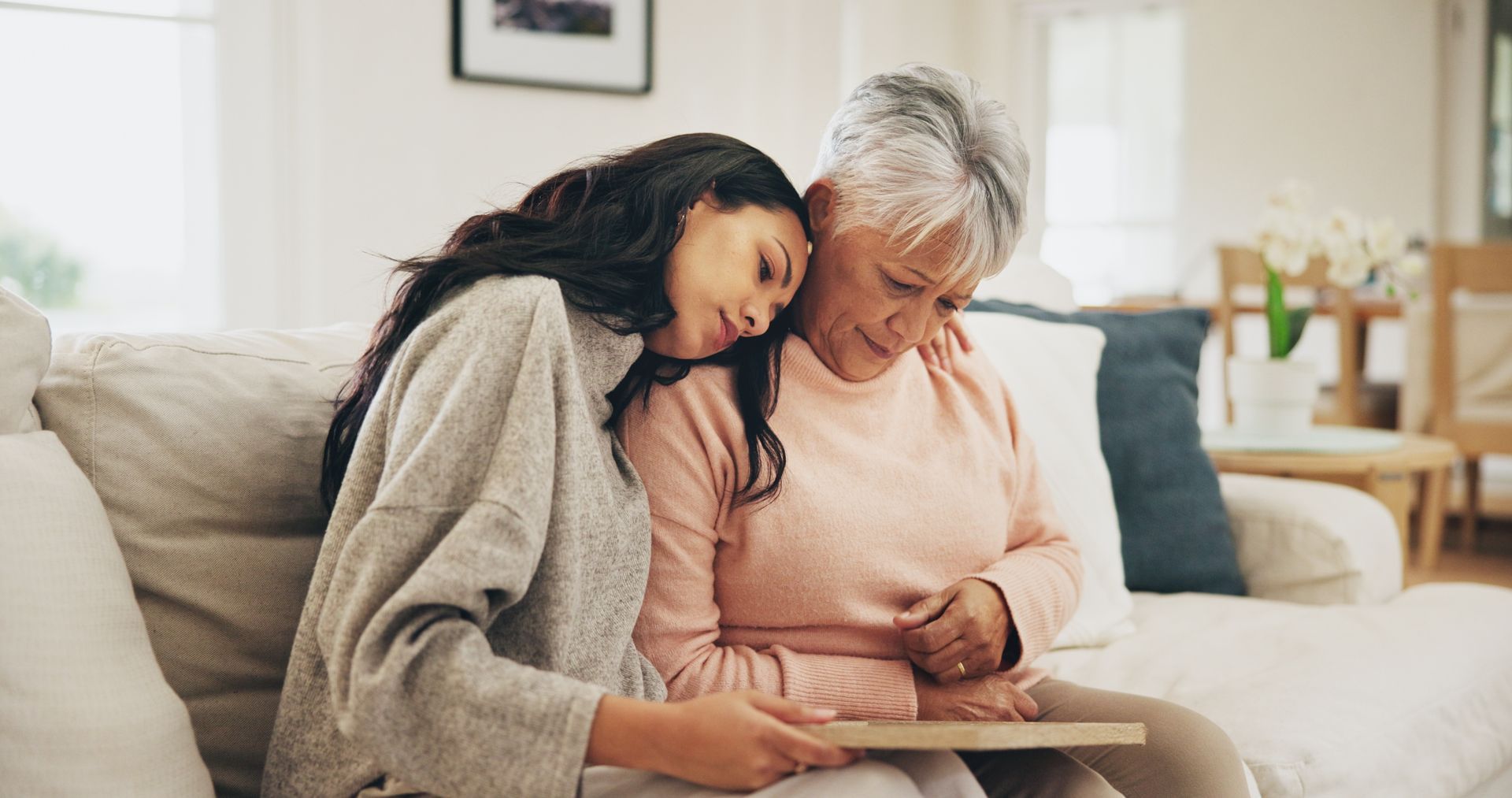 A person leans their head on another's shoulder as they sit on a couch together, looking down at a flat object in their hands.