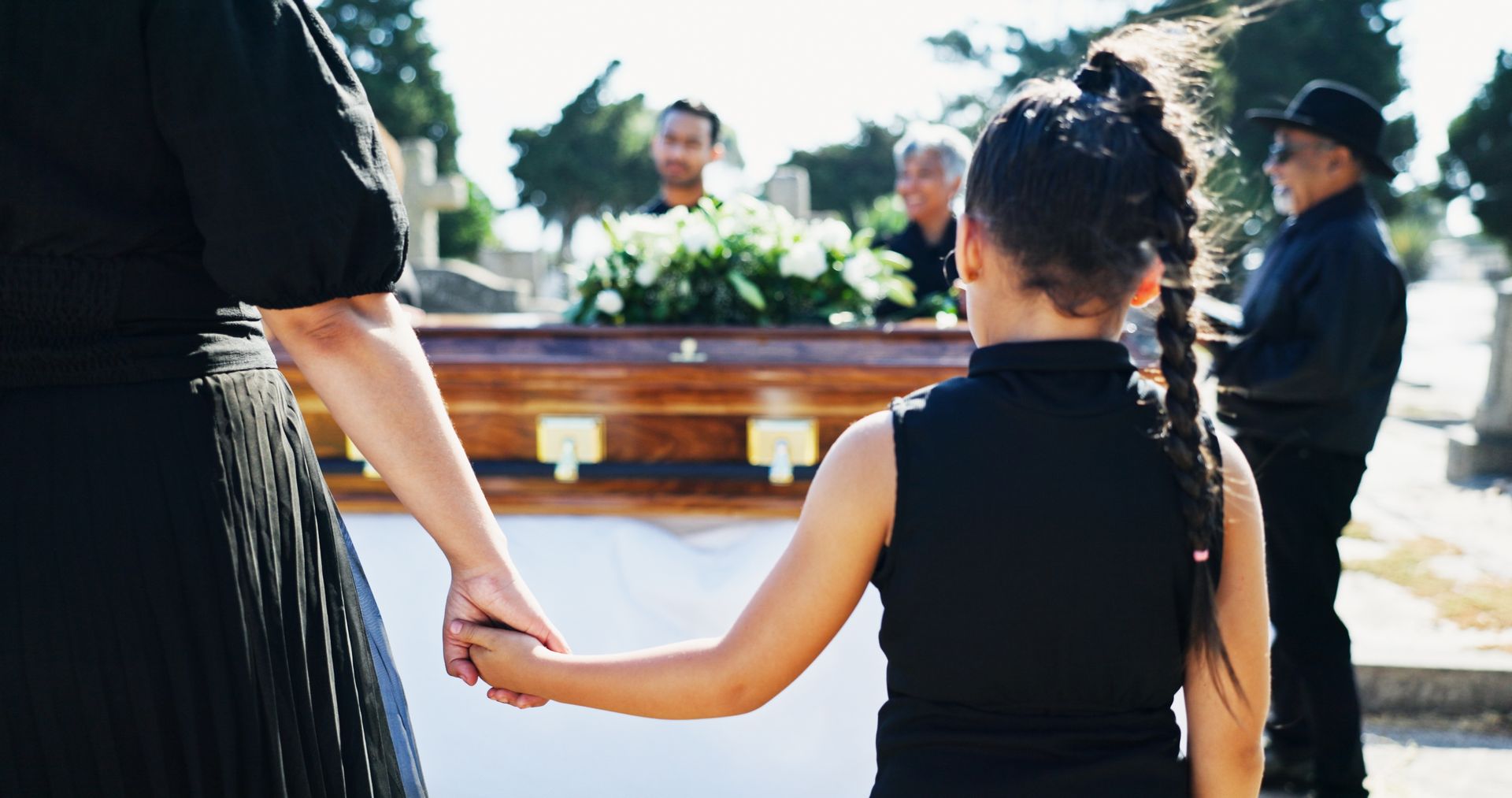 A person and a child in black clothes hold hands while standing before a wooden casket in an outdoor cemetery.