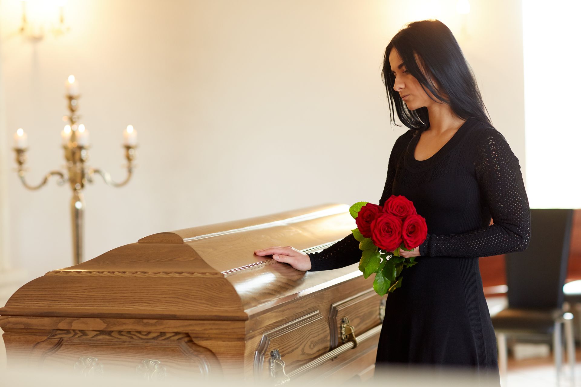 A person in a black dress holds red roses while resting their hand on a wooden casket in a funeral hall.