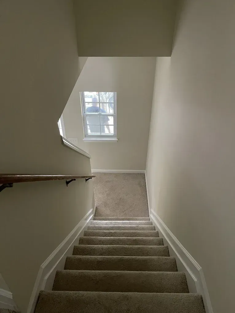 A view looking down carpeted interior stairs toward a small window, with walls painted a neutral off-white.