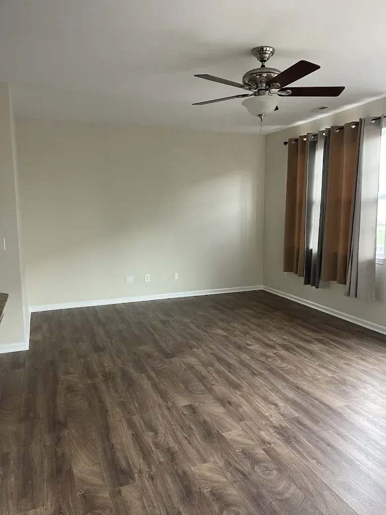 An empty room with neutral walls, dark wood-look plank flooring, a ceiling fan, and windows covered with brown curtains.