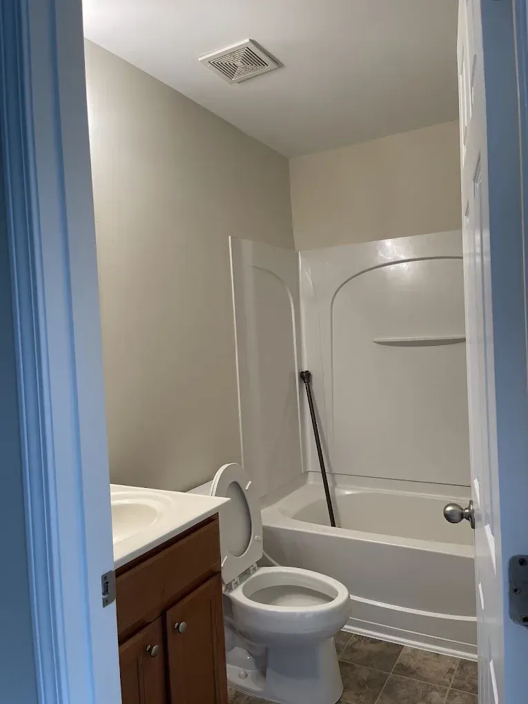 A small, neutral-toned bathroom featuring a wood-toned vanity, a white toilet, and a white bathtub-shower combo.