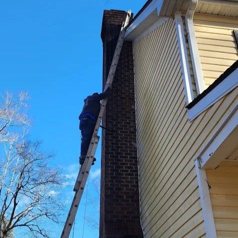 A person climbs an extension ladder leaned against the brick chimney of a yellow-sided house under a clear blue sky.