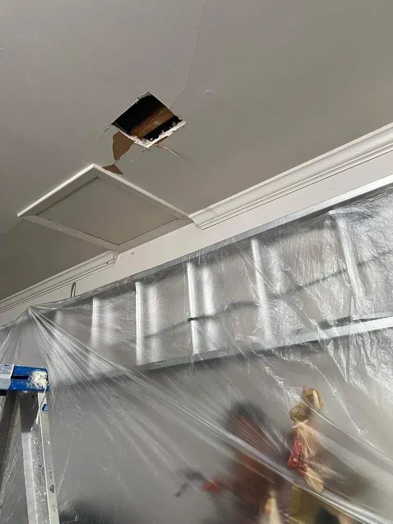 A ceiling repair project in progress, featuring an open hole near a white access hatch and plastic sheeting draped below.
