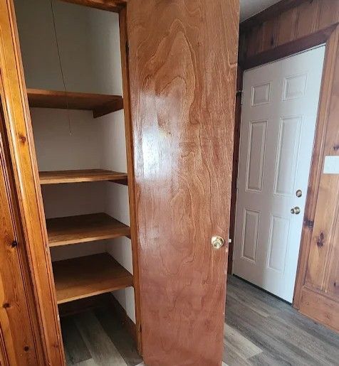 A wooden closet with open shelves next to a white interior door in a room with wood-paneled walls and vinyl flooring.